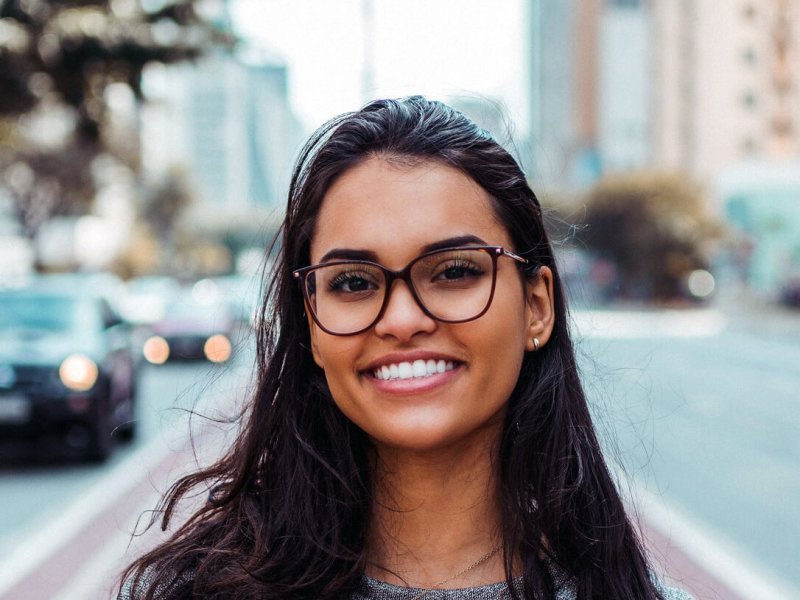 A woman smiling at the camera while standing outdoors near a car and building, wearing stylish clothing and eyewear.