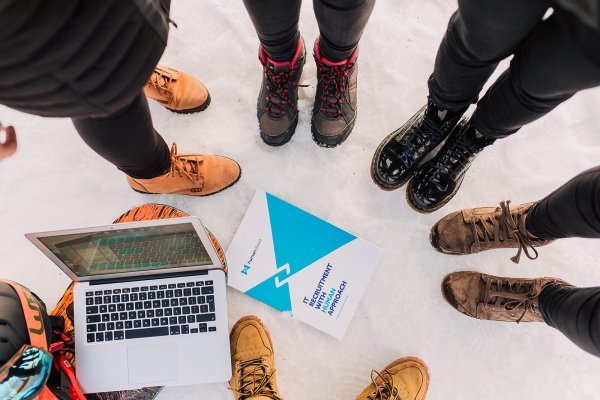 Six people in boots stand in a circle on snow around a laptop and a blue-and-white booklet placed on the ground.