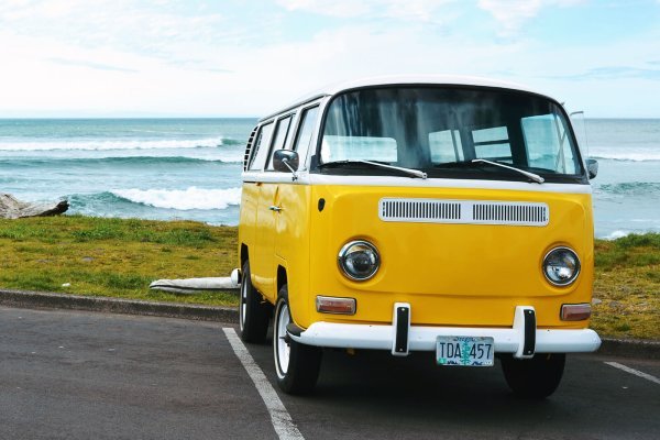 A yellow Volkswagen Type 2 van parked in a parking lot near the beach, with grass and an ocean view in the background.