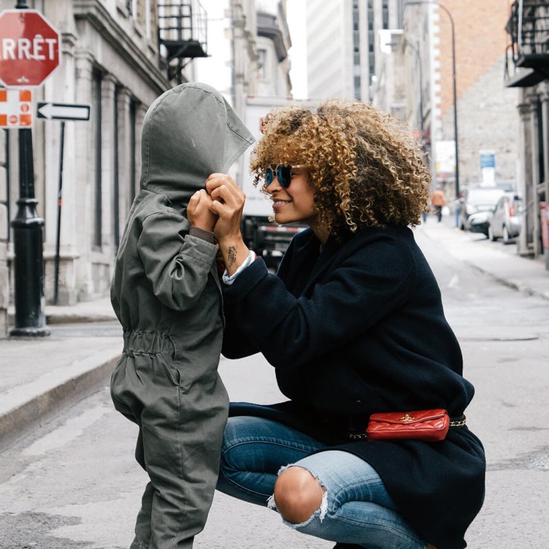 An adult crouches on a city street holding hands with a child in a hooded jumpsuit. Buildings and a stop sign are visible in the background.